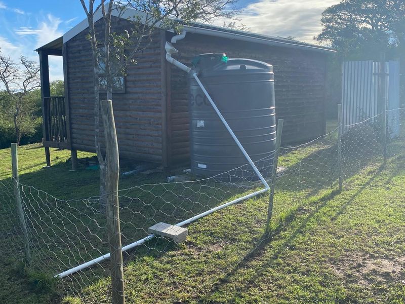 Rear view of a timber cabin featuring a water storage tank, guttering system, and wire mesh perimeter fencing on a grassy plot.