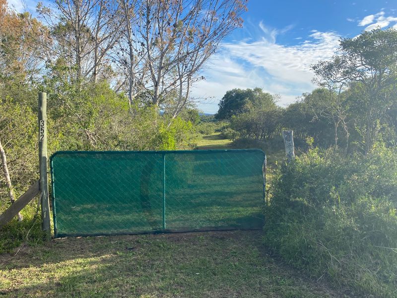 A view of the entrance to a vacant residential plot, featuring a green mesh gate and natural vegetation.