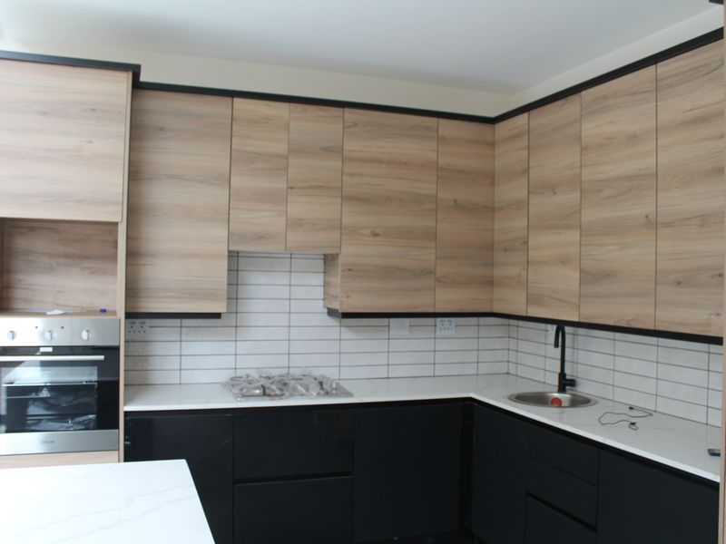 Modern kitchen featuring wood-grain upper cabinets, matte black lower cabinets, white stone countertops, subway tile backsplash, and integrated stainless steel oven and gas hob.