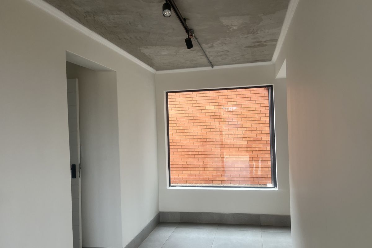 A bright, empty room featuring an exposed concrete ceiling, modern track lighting, large picture window, and neutral tiled flooring.