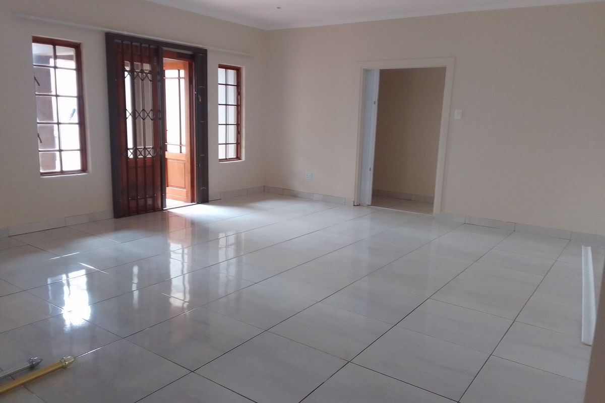 A spacious, empty living room featuring neutral walls, polished tiled flooring, and timber-framed windows with an integrated security gate leading to the exterior.