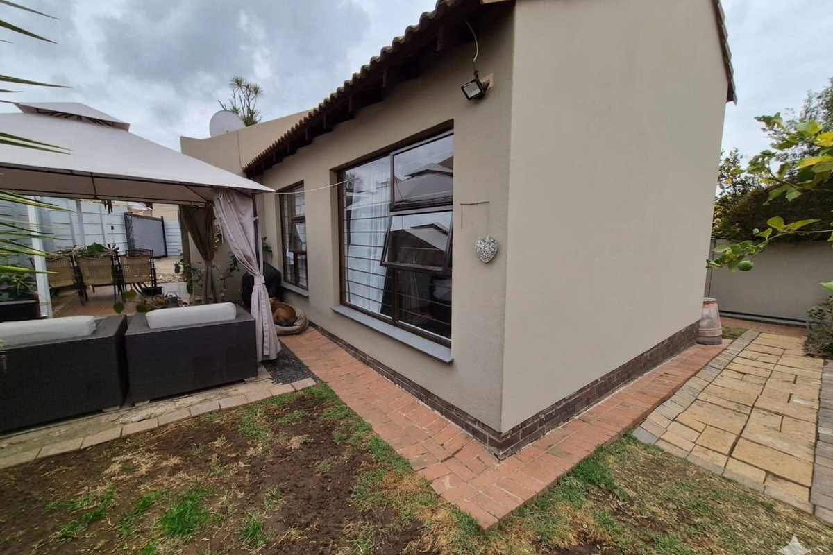 Rear view of the property featuring plastered exterior walls, aluminium window frames, and brick paved pathways leading to a covered patio area.