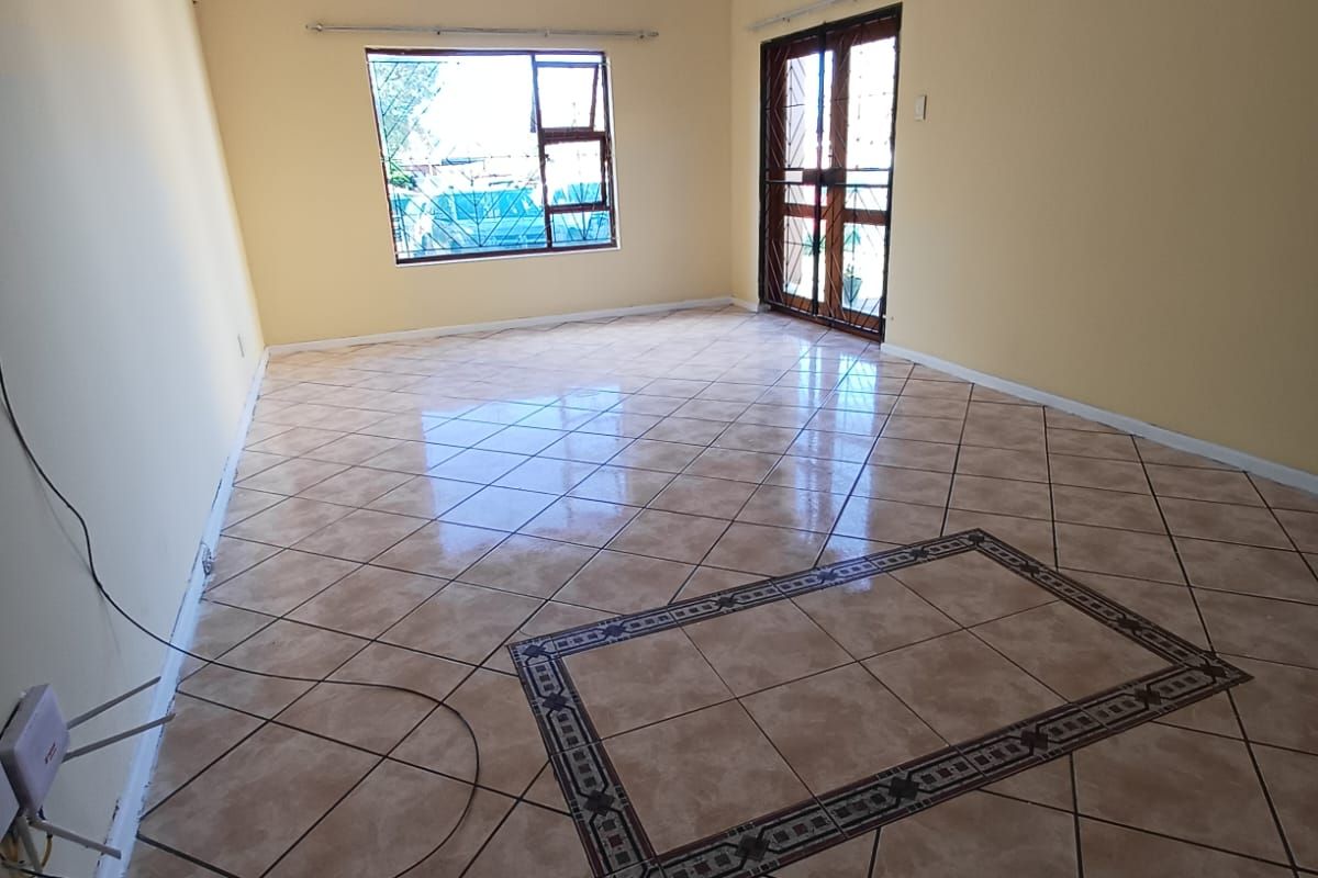 A spacious, empty living area featuring neutral wall paint, tiled flooring with a decorative inlay, and access to an exterior door with a security gate.