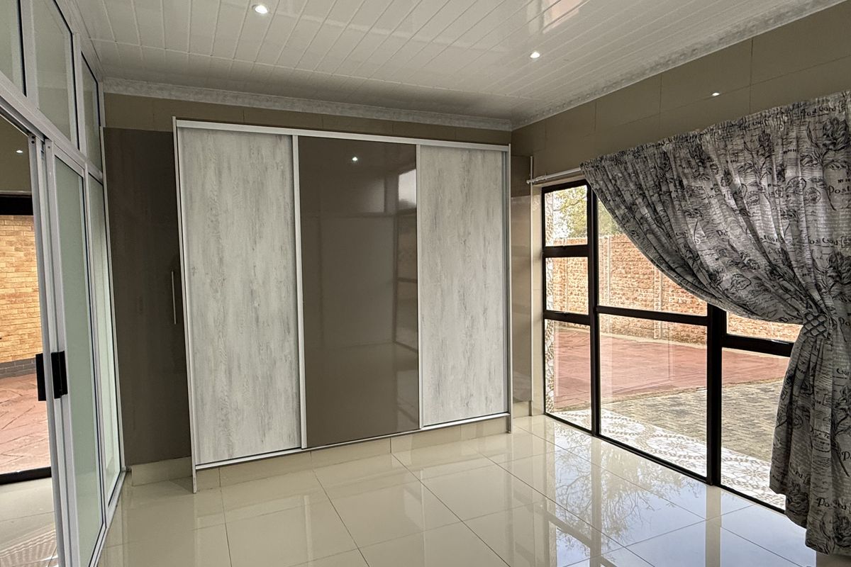 A bright, empty bedroom featuring modern built-in sliding door wardrobes, polished tile flooring, and large aluminium-framed windows with access to the exterior.