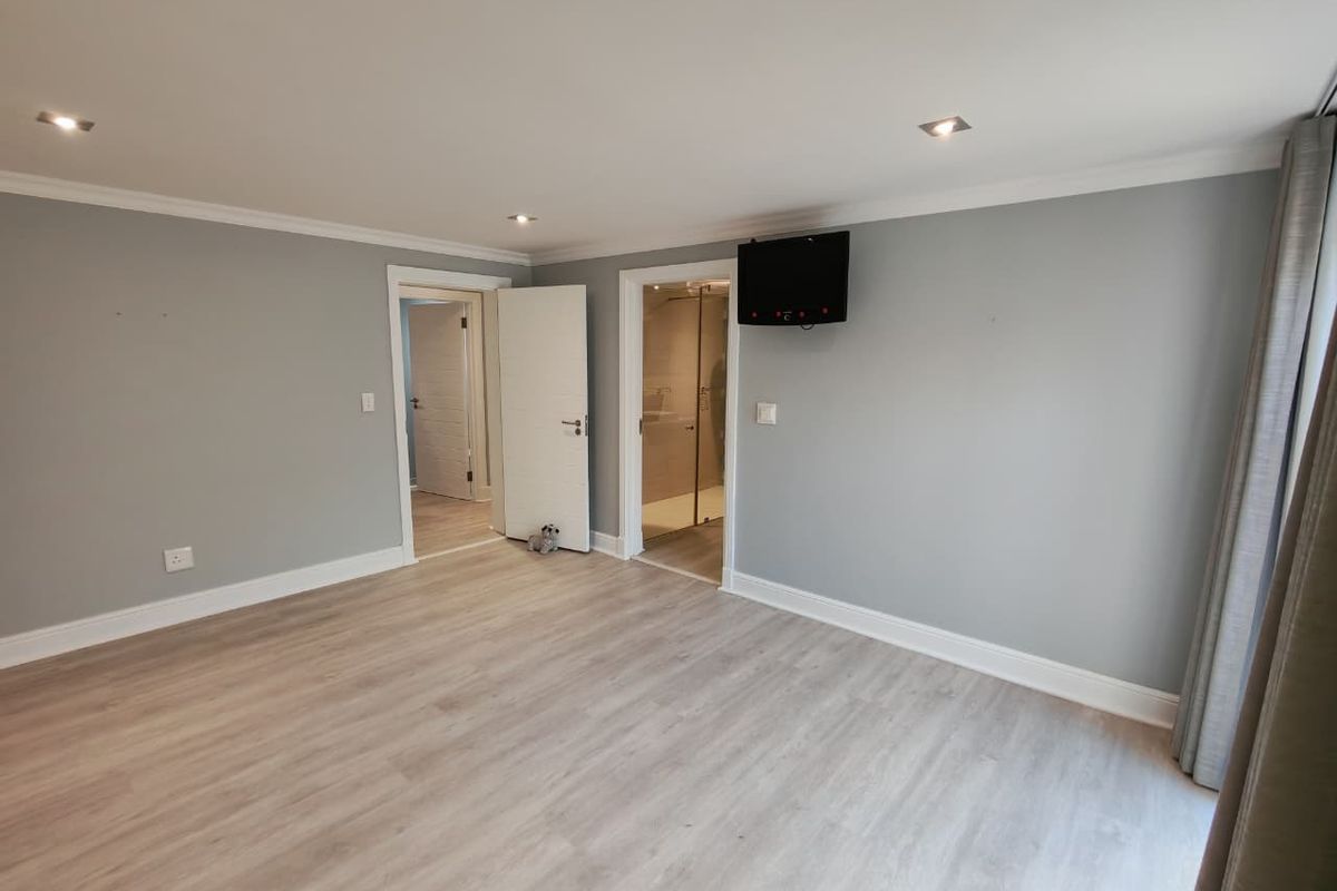 A spacious bedroom featuring light wood-look flooring, recessed ceiling lighting, and an en-suite bathroom entrance.