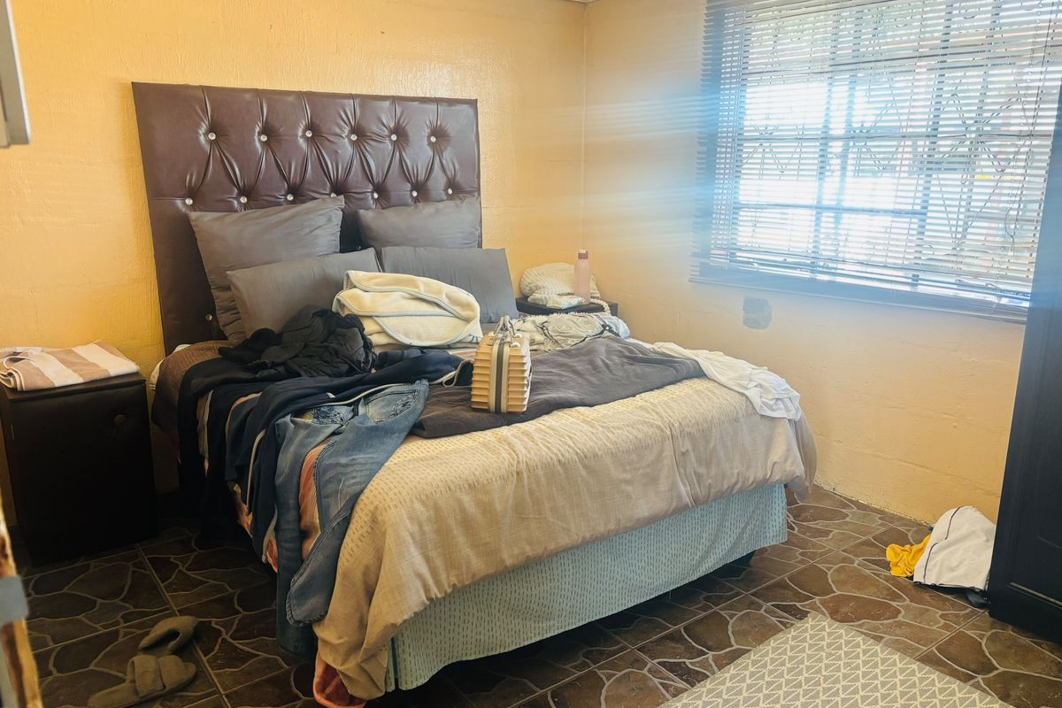 A bedroom featuring tiled flooring, a window with fitted blinds, and a tufted headboard. The room requires cosmetic updates to improve its overall condition.