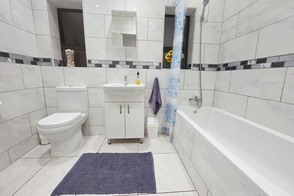 A clean, modern bathroom featuring white marble-look wall and floor tiles, a bathtub with shower attachment, and a vanity unit with a basin.