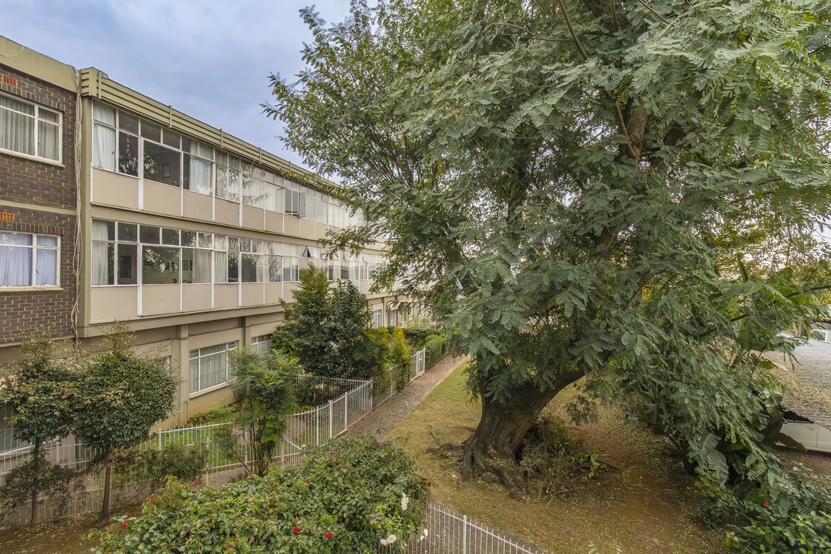 Exterior view of the apartment building facade featuring large windows, brickwork, and a mature garden area with established trees.