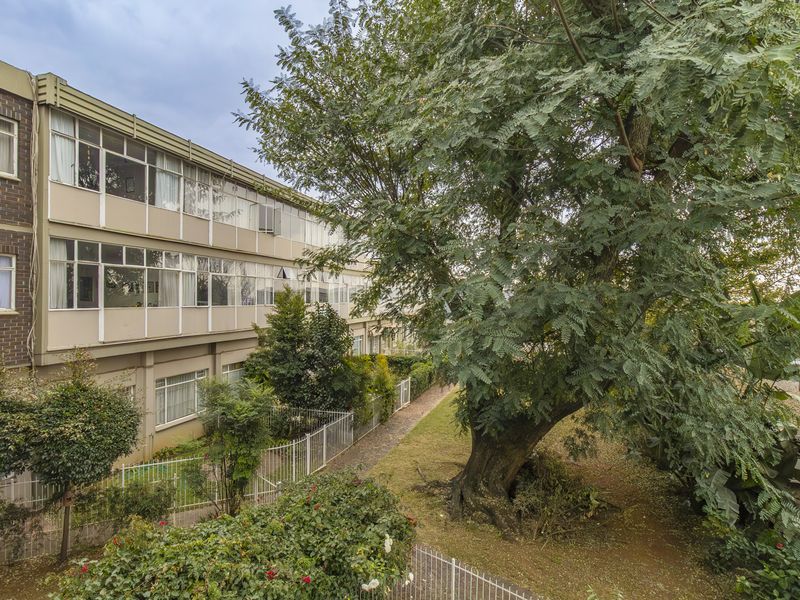 Exterior view of the apartment building facade featuring large windows, brickwork, and a mature garden area with established trees.