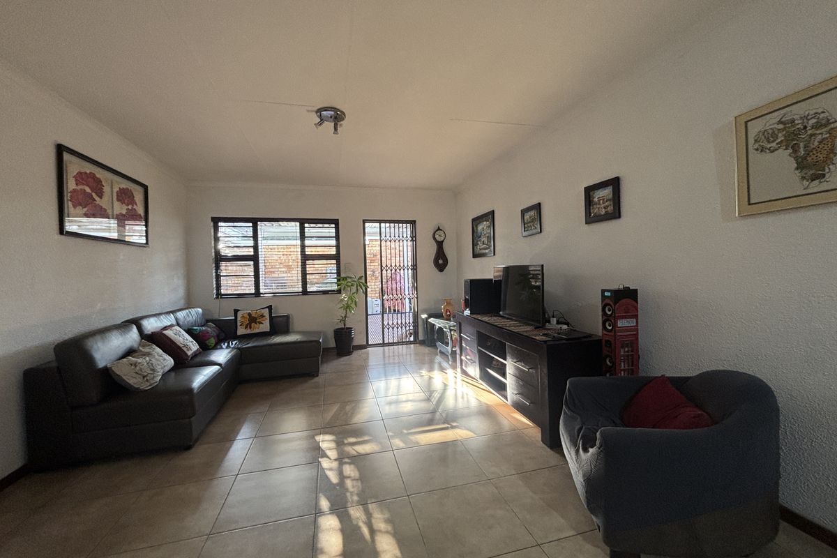 A spacious living room featuring tiled flooring, natural light from the window, and a secure entrance door with a metal security gate.