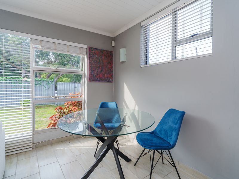 A bright dining area featuring tiled flooring, large windows with venetian blinds, and a clean, neutral aesthetic overlooking the garden.
