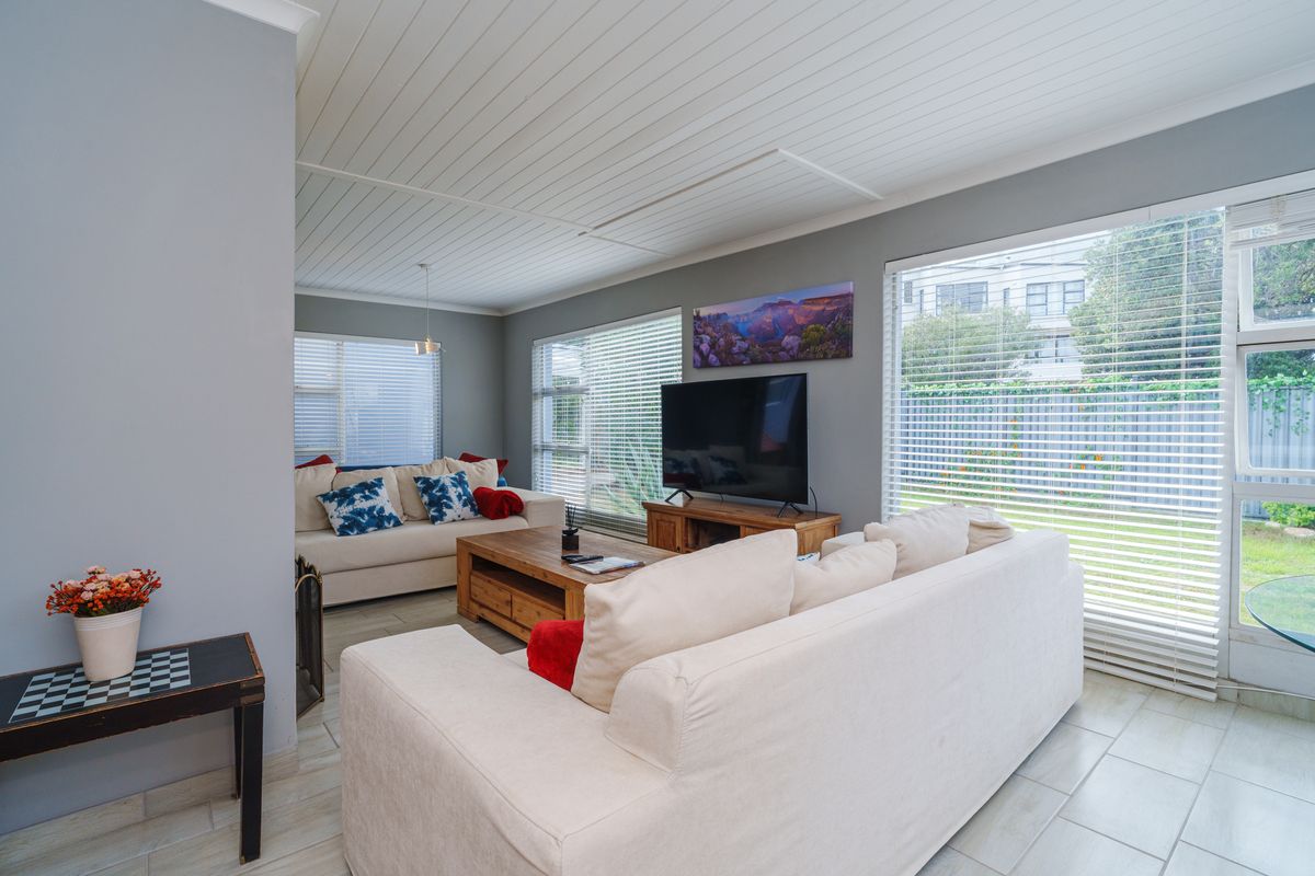A bright and airy living room featuring tiled flooring, a white tongue and groove ceiling, and large windows fitted with venetian blinds.