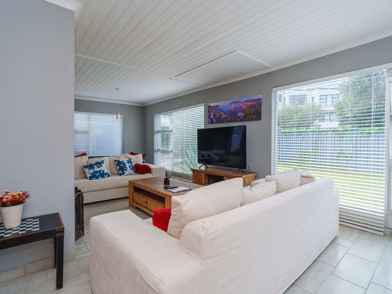 A bright and airy living room featuring tiled flooring, a white tongue and groove ceiling, and large windows fitted with venetian blinds.