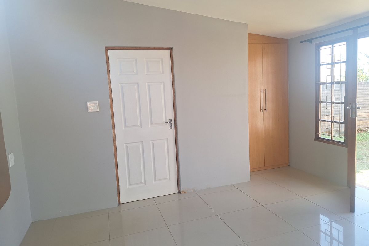 A bright bedroom featuring neutral grey walls, light-colored floor tiles, and a built-in wooden wardrobe with a window overlooking the garden.
