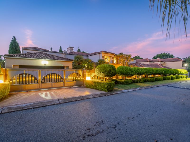 A well-maintained cluster home exterior featuring a secure metal gate, paved driveway, and manicured garden hedges at dusk.