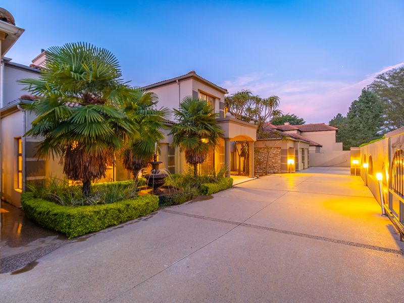 A well-maintained front exterior view of a cluster property featuring a paved driveway, landscaped garden beds, a water fountain, and modern exterior lighting.