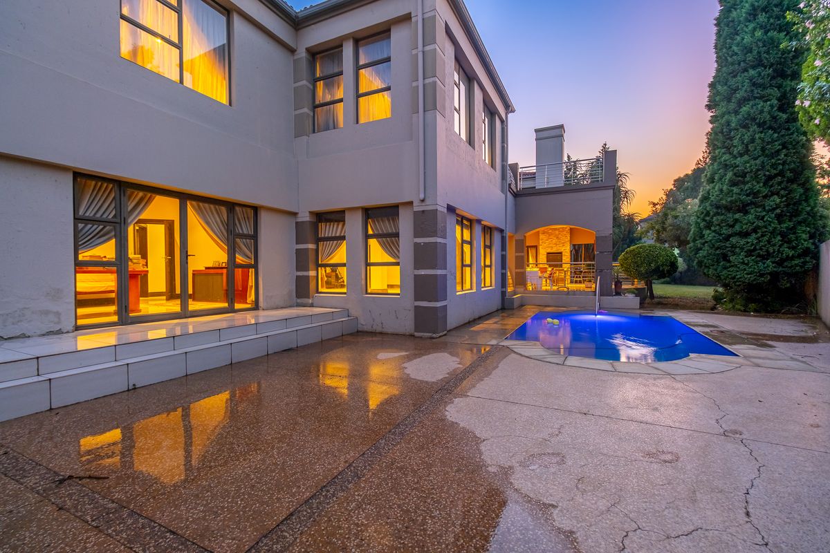 Rear exterior view of the property featuring a swimming pool, covered patio with built-in braai, and a spacious paved entertainment area at dusk.