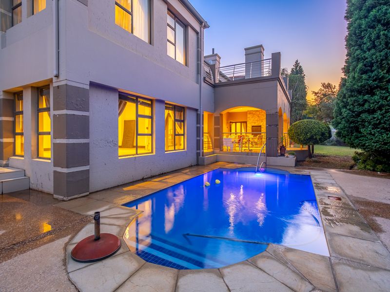 A well-maintained swimming pool area featuring stone paving, integrated pool lighting, and a covered patio with an upper-level balcony at dusk.
