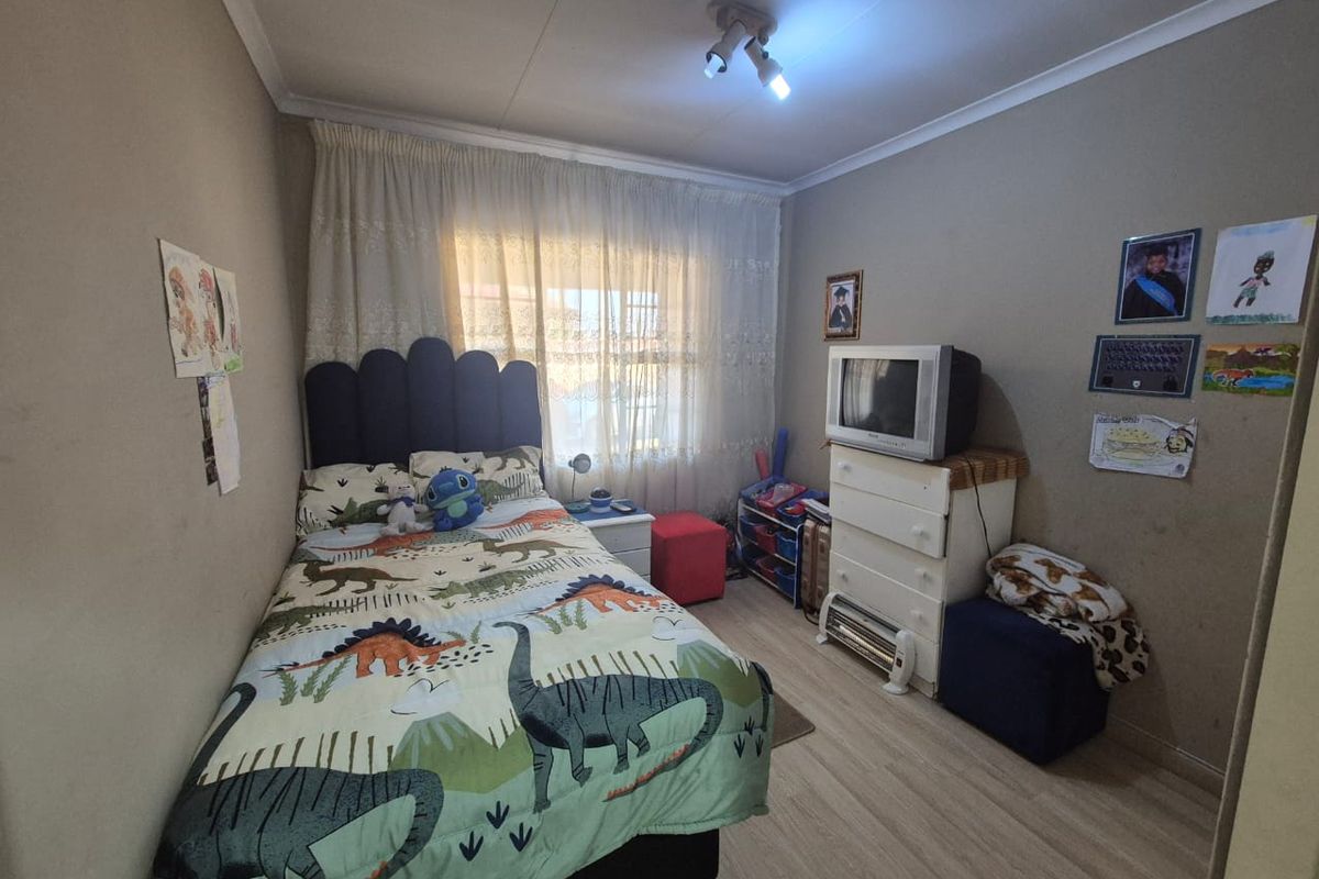 A functional bedroom featuring light-toned laminate flooring, neutral wall finishes, and a window with sheer curtains providing natural light.