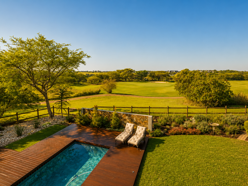 A beautifully maintained outdoor pool area featuring timber decking, a stone retaining wall, and a lush garden overlooking a scenic golf course landscape.