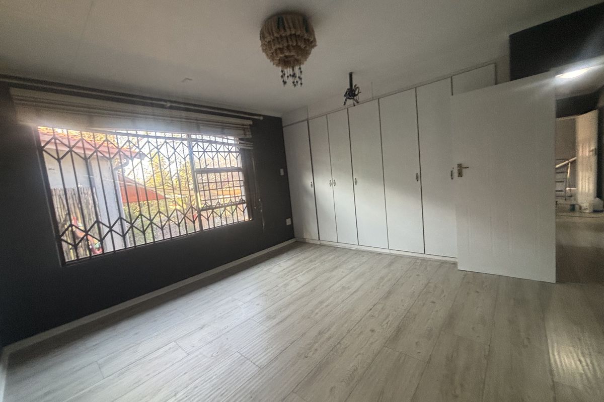 A bright, empty bedroom featuring light-toned laminate flooring, extensive built-in white cupboards, and security bars on the window.