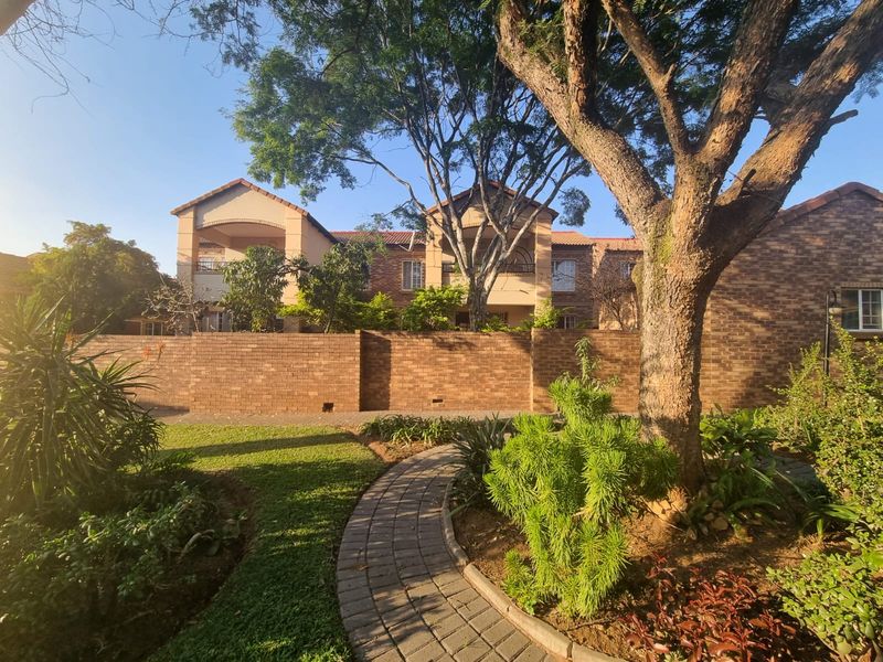 Rear view of the townhouse complex featuring a brick boundary wall, paved garden pathway, and established trees under a clear sky.