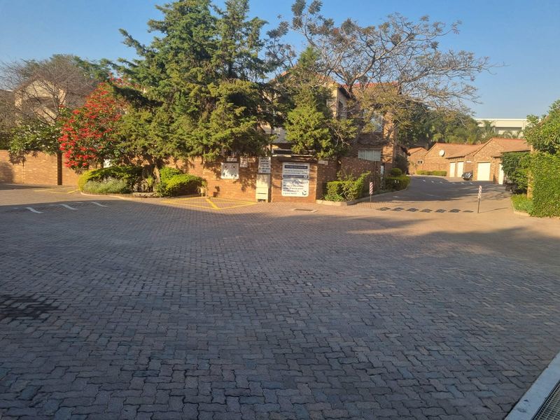 View of the paved entrance and driveway leading to the townhouse complex, featuring established trees and brick building facades.