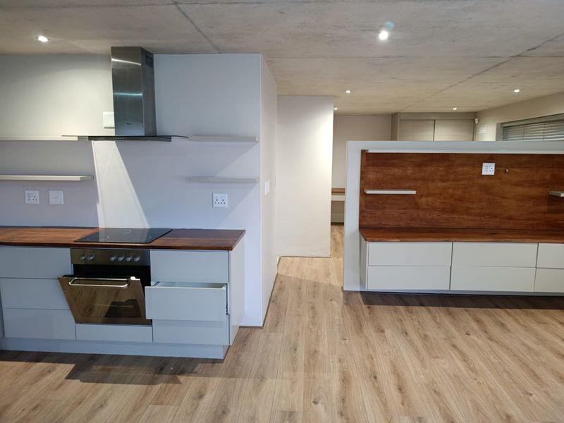 Modern open-plan kitchen and living area featuring laminate flooring, exposed concrete ceilings, built-in cabinetry, and integrated cooking appliances.