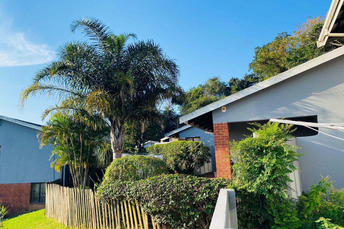 Exterior view of the apartment building featuring a covered patio area, brick pillar accents, and established garden landscaping with mature palm trees.