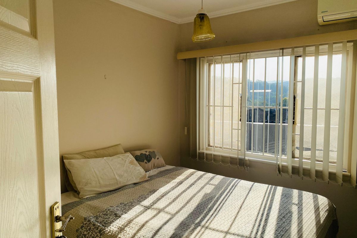 A cozy bedroom featuring a wall-mounted air conditioning unit, vertical window blinds, and security bars on the window.