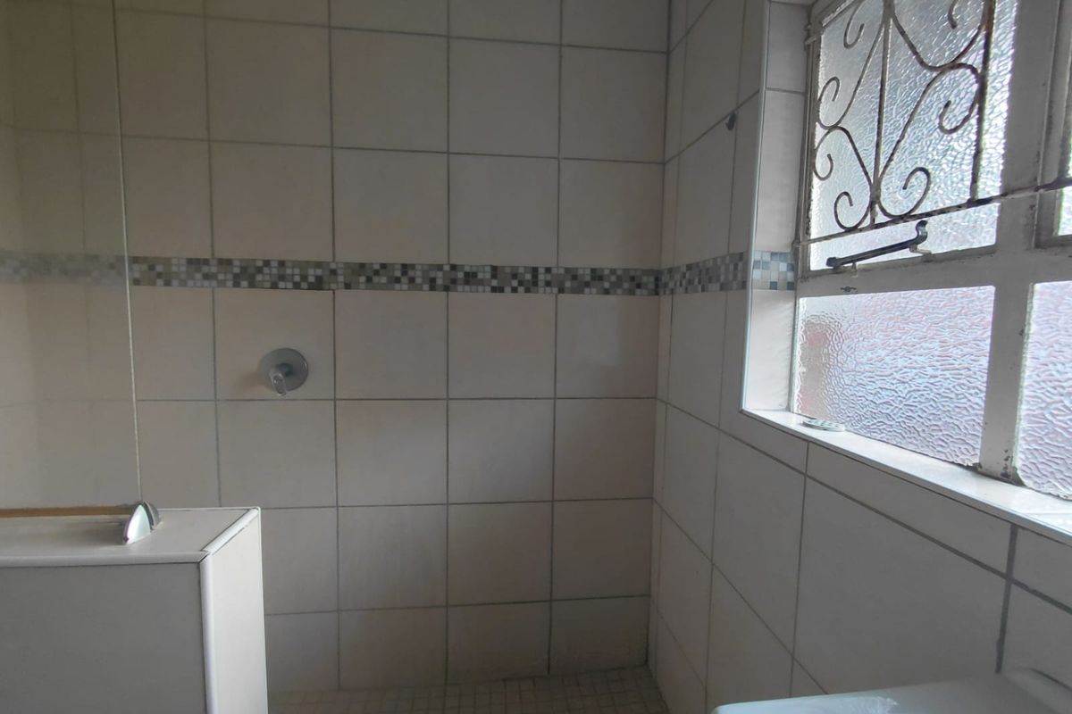 A functional bathroom shower area featuring tiled walls with a mosaic accent border and a window with security bars.
