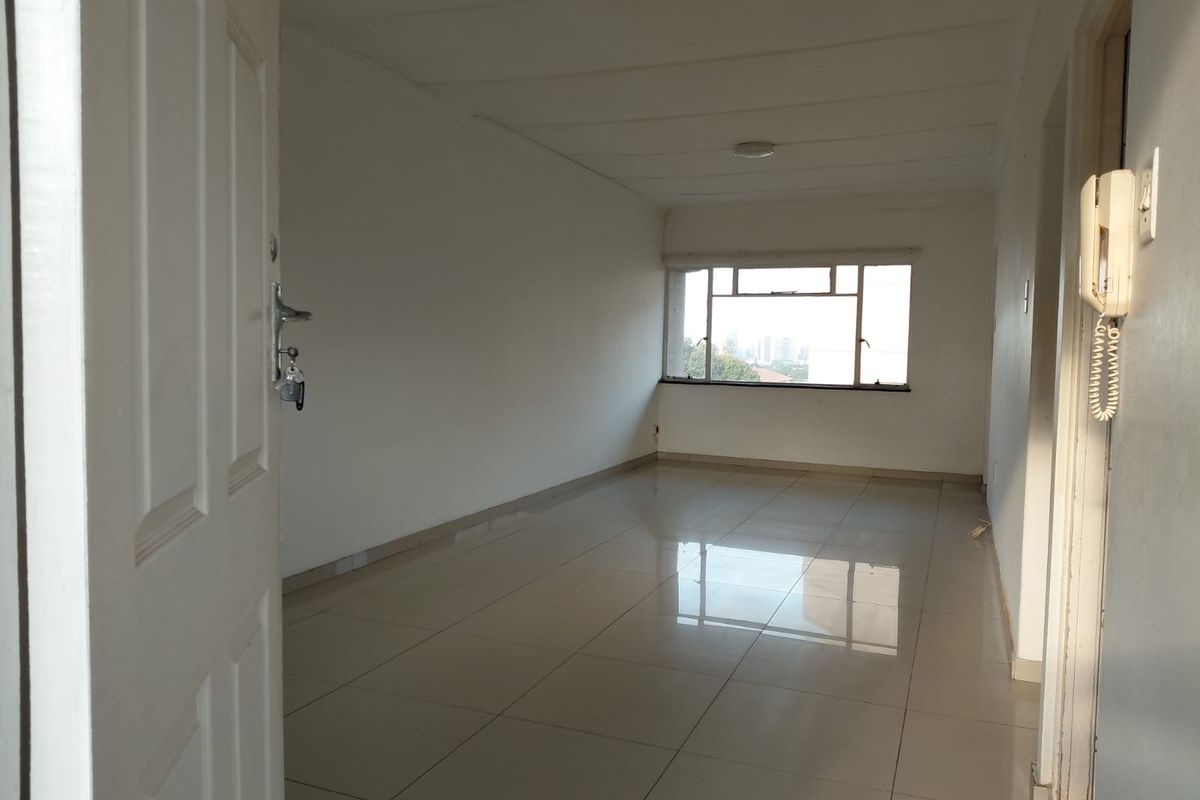 An empty, open-plan living area featuring neutral tiled flooring, white walls, and a large window providing natural light.