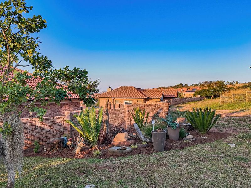 A well-maintained garden area featuring landscaped beds, brick boundary walls, and views of neighboring residential properties under a clear blue sky.
