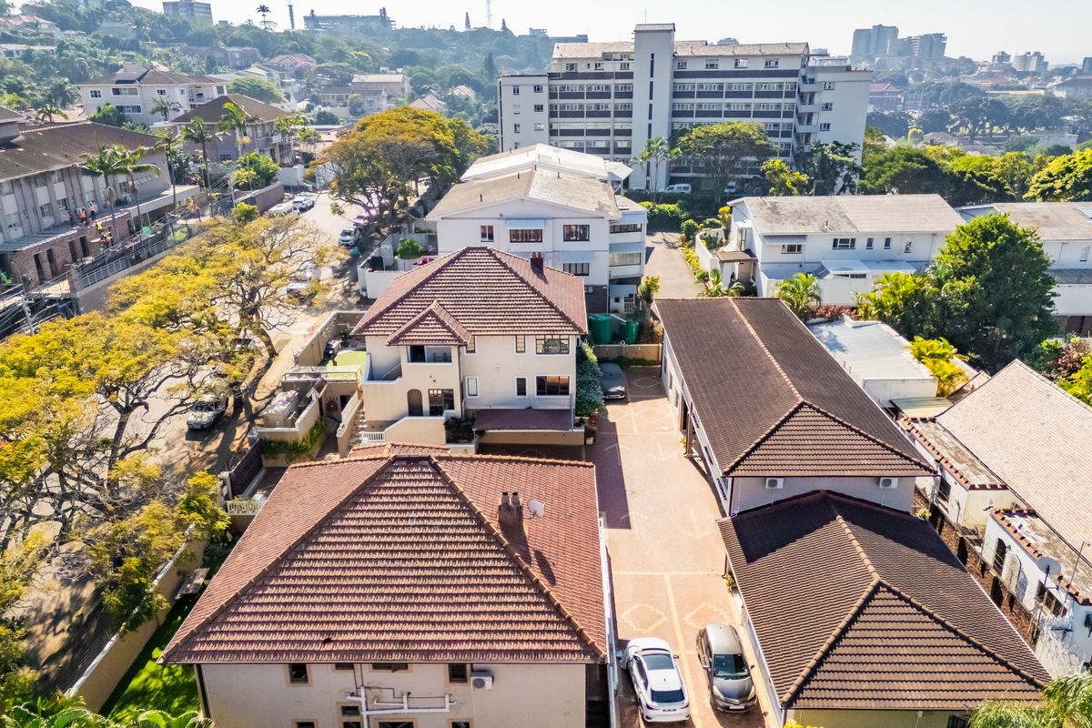 Aerial view of the apartment complex showing multiple buildings with tiled roofs, paved driveway, and surrounding urban landscape.