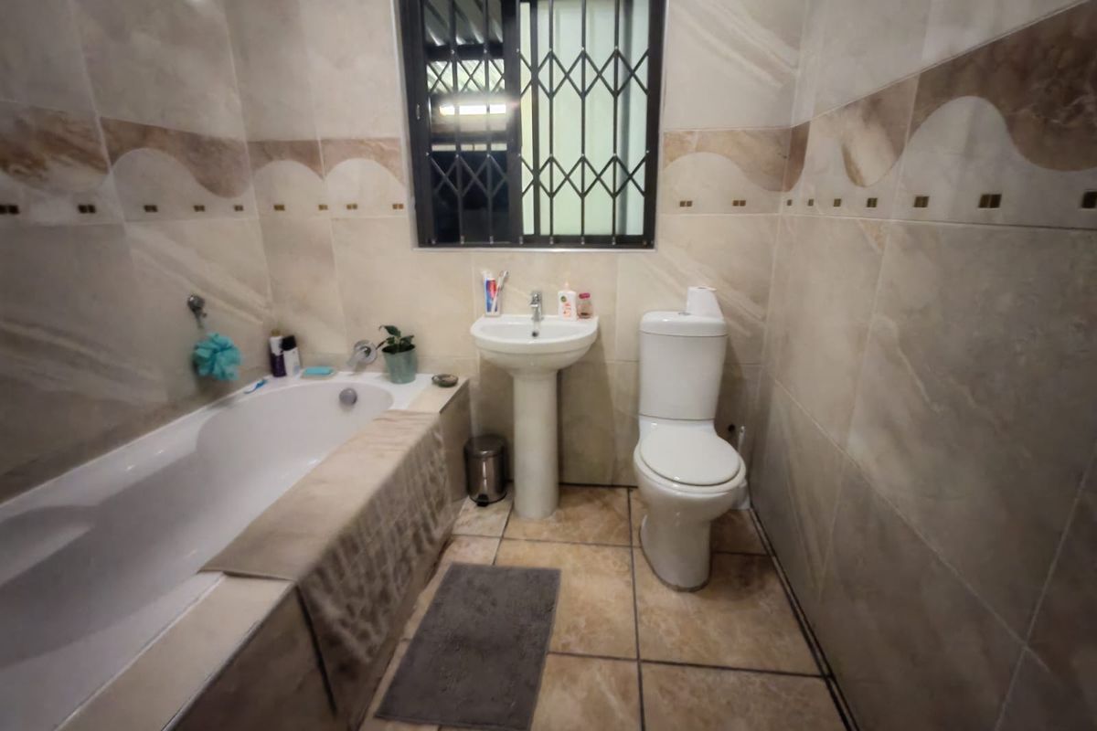 A functional bathroom featuring a bathtub, pedestal basin, and toilet, finished with neutral wall and floor tiling and a security-grille window.