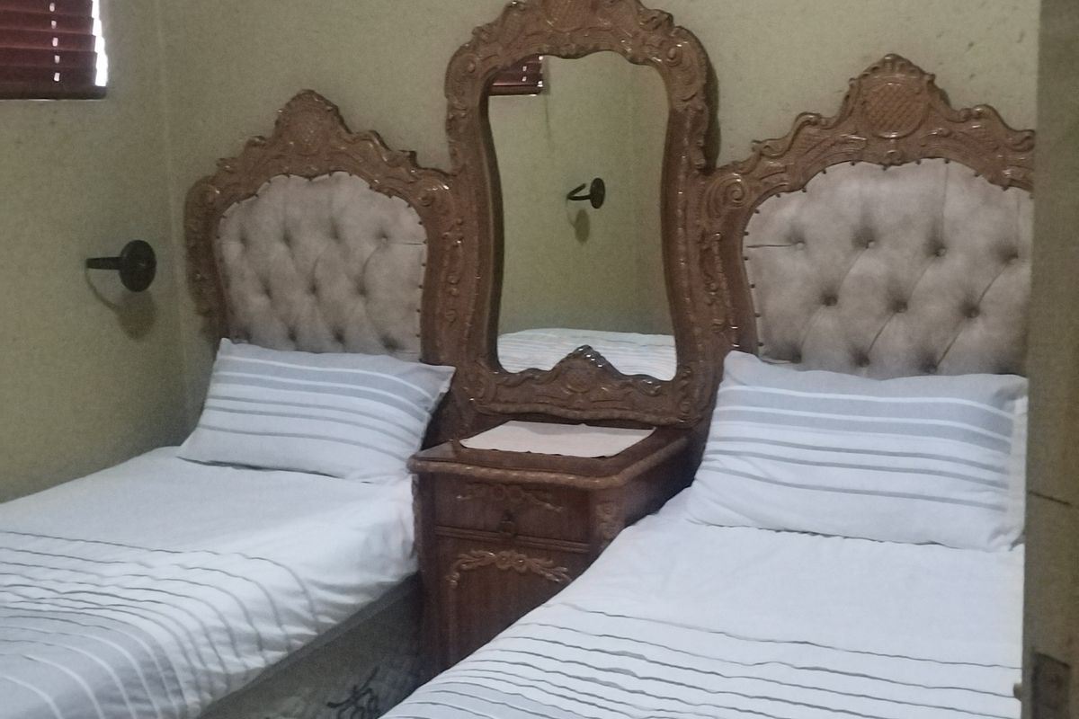 A bedroom featuring a central built-in vanity mirror and headboard unit between two bed spaces, with wooden window blinds.