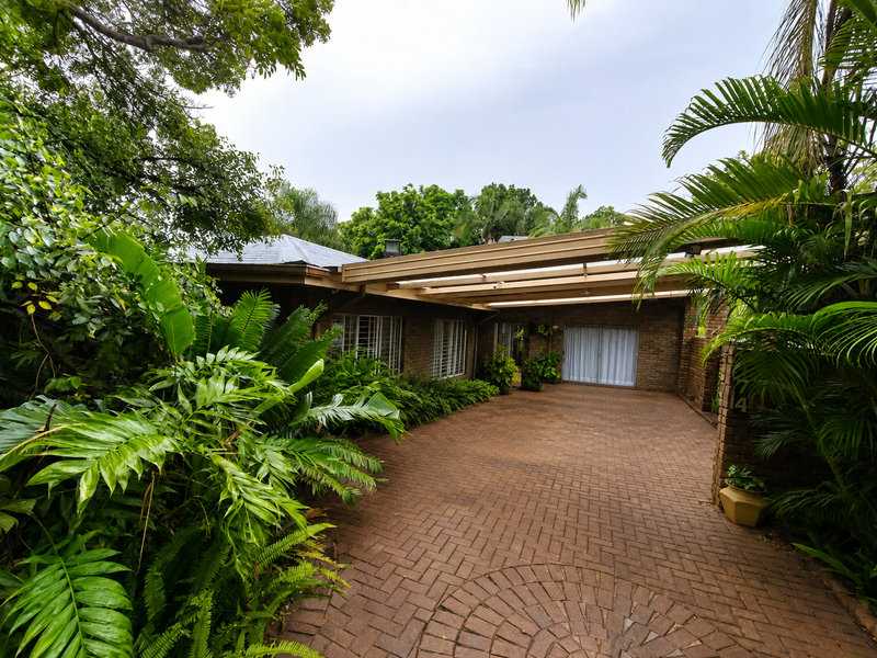 Front exterior view featuring a brick driveway, covered carport, and established garden surroundings leading to the main entrance.