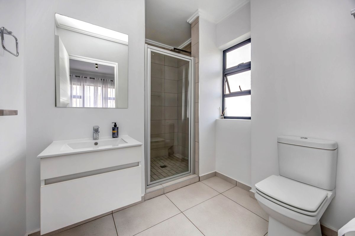 A clean, modern bathroom featuring a white vanity with basin, a glass-enclosed shower, and a standard toilet with neutral floor tiling.