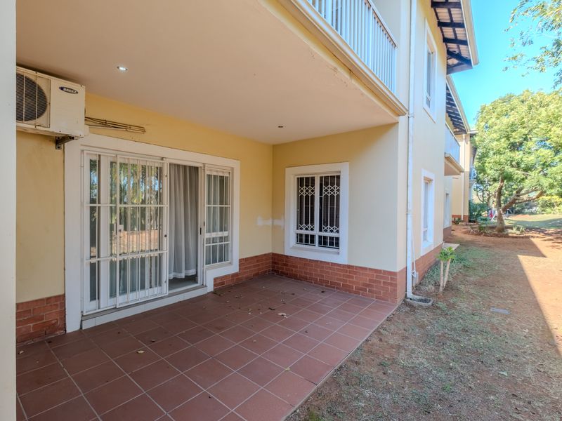A covered ground-floor patio featuring tiled flooring, security bars on windows, and a sliding door entrance, overlooking a garden area.
