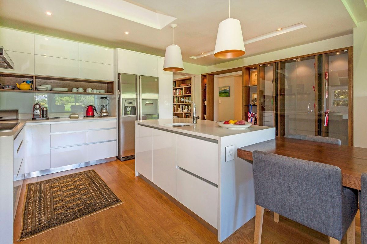 Modern kitchen featuring high-gloss white cabinetry, stone countertops, a central island with sink, and warm hardwood flooring, complemented by stylish pendant lighting.
