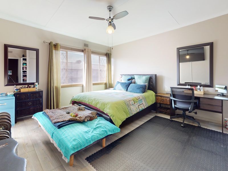 A well-lit bedroom featuring laminate flooring, a ceiling fan, and large windows with venetian blinds, offering a functional and comfortable living space.