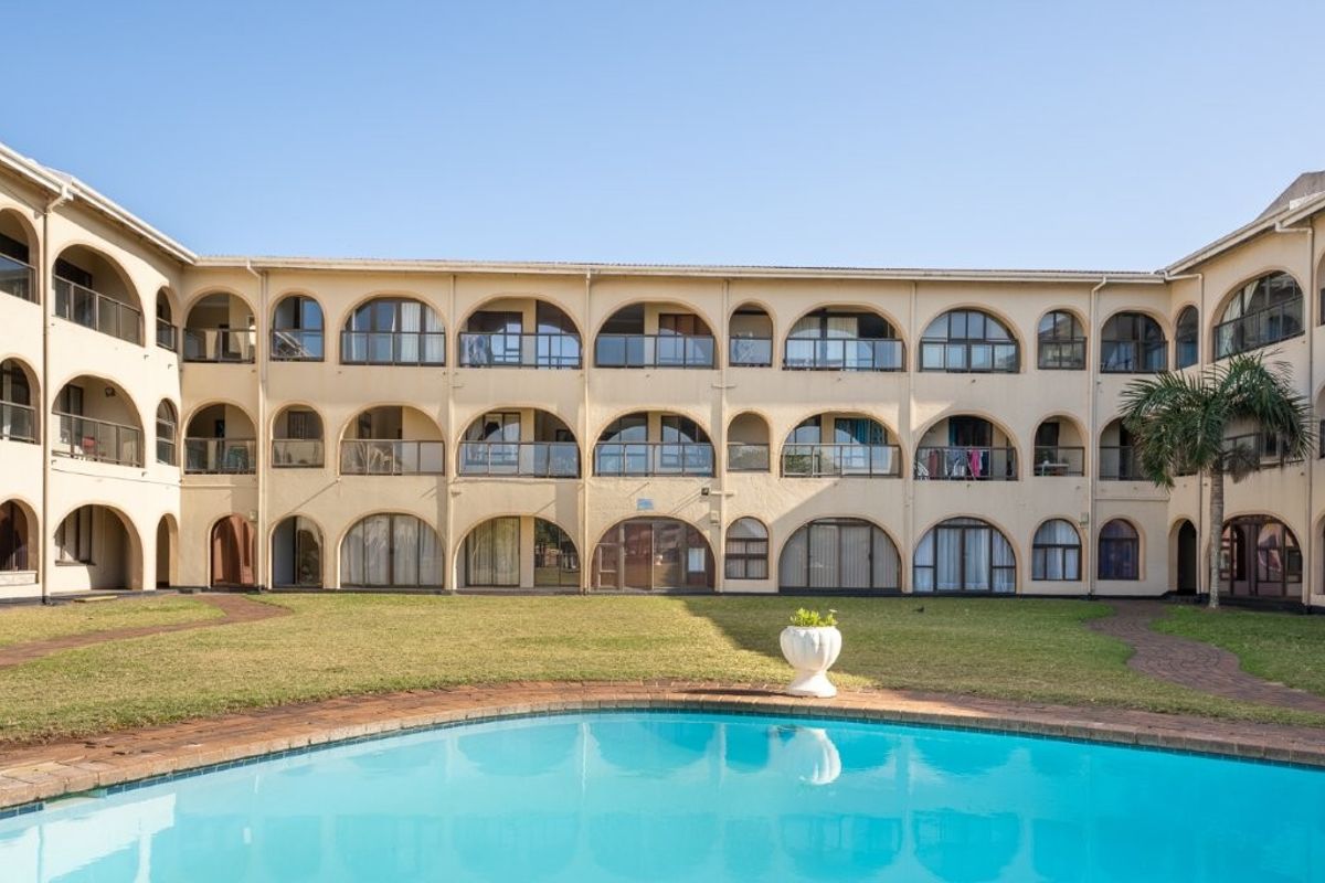 Exterior view of an apartment complex featuring a central swimming pool, manicured lawn, and multiple levels of arched balconies with glass railings.