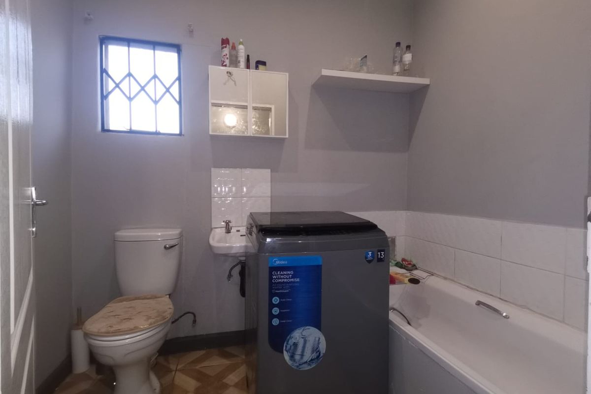 A functional bathroom featuring a bathtub, toilet, and small washbasin, with wall-mounted storage and a security-gated window.