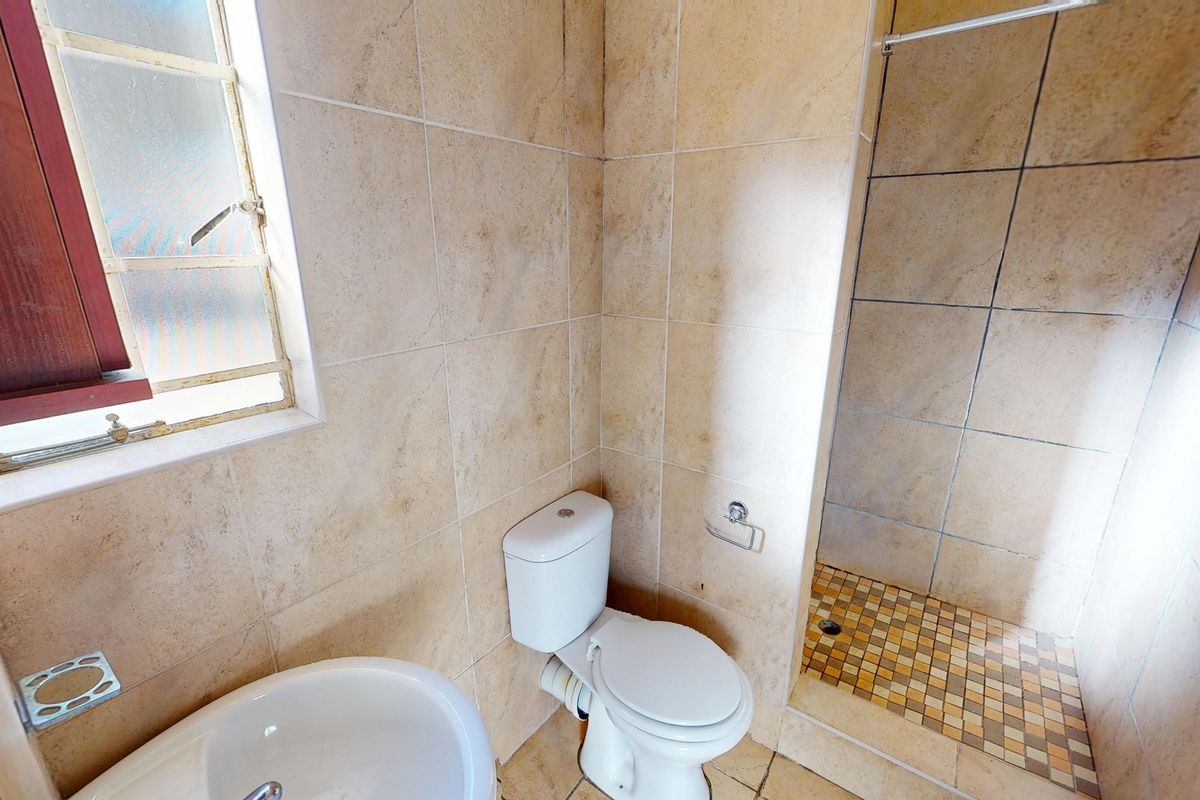 A functional bathroom featuring a shower enclosure, pedestal basin, and toilet with neutral ceramic wall tiling and mosaic floor accents.