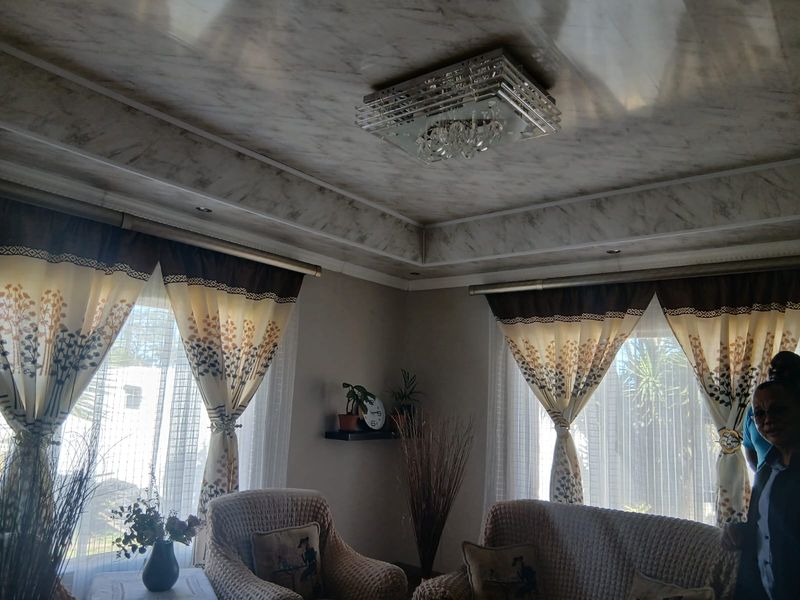A living room featuring a decorative tray ceiling with recessed lighting and a central crystal chandelier, complemented by large windows with curtain fixtures.