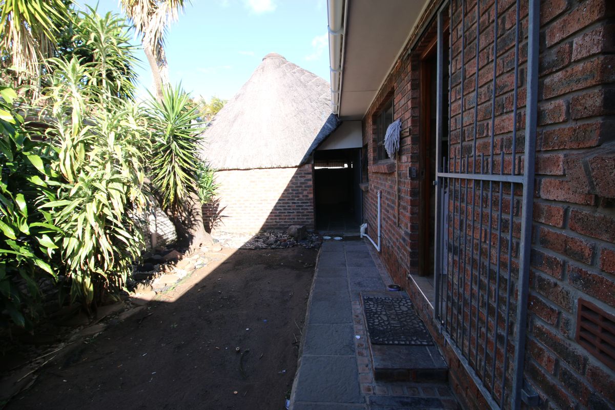 A rear exterior view showing a brick facade, a thatched roof structure, and a paved walkway leading along the side of the house.