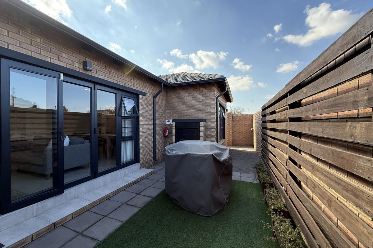 A well-maintained rear courtyard featuring a paved patio, artificial lawn, built-in braai, and modern aluminium sliding doors leading into the living area.