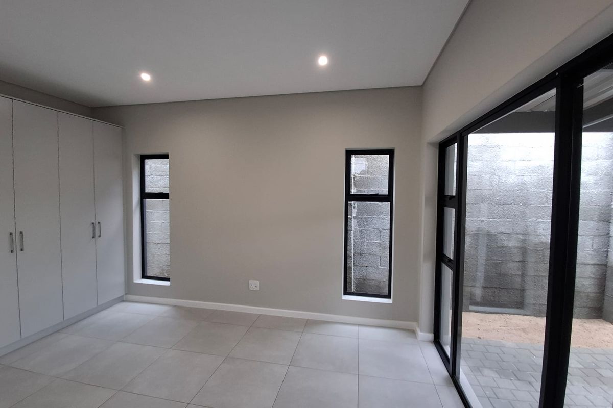 A modern, neutral-toned bedroom featuring built-in cupboards, large format floor tiles, and sliding glass doors providing access to an outdoor area.