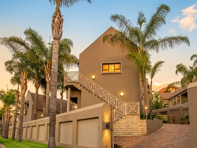 Exterior view of a modern cluster home featuring a paved driveway, external staircase with metal railings, and landscaped palm trees.