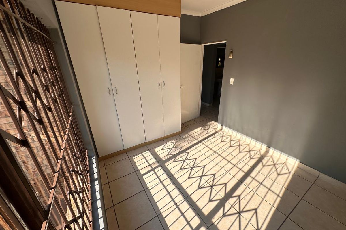 A bright, empty bedroom featuring tiled flooring and a built-in wardrobe, secured with a metal security gate on the window.